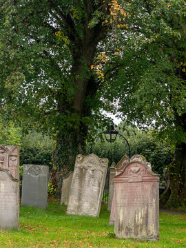 27th September 2026, Embleton, Cumbria. Ancient weathered gravestones stand beneath the lush green canopy of mature trees in a tranquil churchyard, evoking a sense of history and solemn peace.