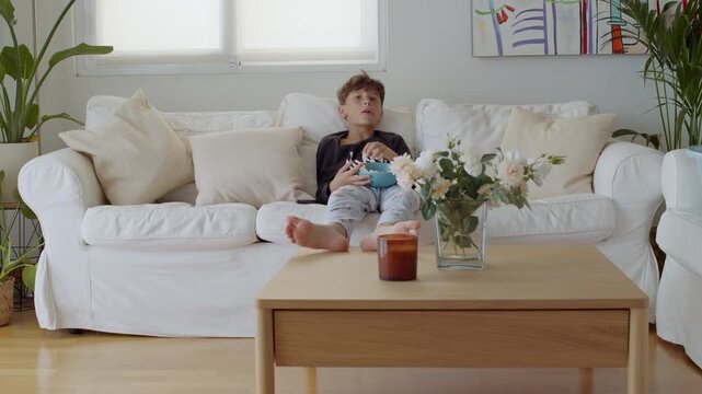 Barefooted boy in pajamas relaxing on a comfortable white sofa in a modern living room, eating popcorn from a bowl while watching an entertaining movie or cartoon on television