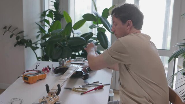 Male luthier performing stepwise electric guitar setup and stringing over workshop workbench