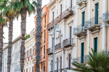 Row of pastel Mediterranean apartment buildings with wrought iron balconies and green shutters behind tall palm trees and a street lamp, concept of southern European city living, real estate and summ © daily_creativity