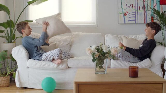 Two young brothers laughing and playing together in a bright living room, joyfully throwing a pillow back and forth while sitting on a comfortable white sofa during a playful morning at home