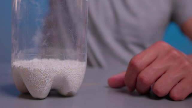 Hand tilting clear plastic bottle while white granules slide and settle across smooth tabletop beside second bottle in soft neutral light, creating simple product handling scene