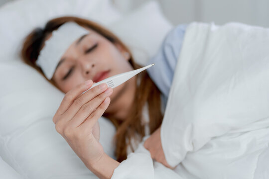 Sick young Asian woman lying in bed with a cooling patch on her forehead, looking at a thermometer while resting under a blanket at home, fever, illness, temperature check, recovery