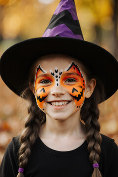 Smiling girl in a witch hat with pumpkin face paint in an autumn park