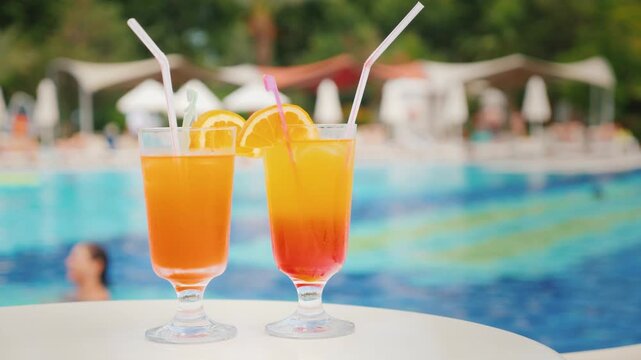 Two tropical orange cocktails with straws standing on white table near turquoise swimming pool of luxury resort during summer vacation.