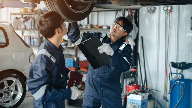 Two Asian male auto mechanics in blue uniform inspecting car underbody with flashlight and checklist at repair shop garage. Auto maintenance, vehicle inspection, and service concept.