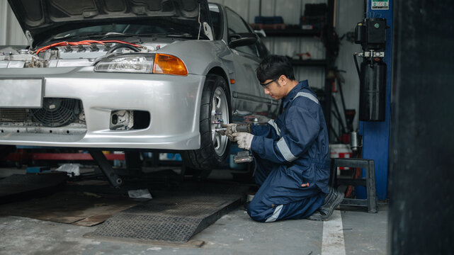 Asian male auto mechanic in blue uniform using cordless impact wrench on car wheel at repair shop garage. Auto maintenance, tire service, brake inspection, and vehicle repair concept.