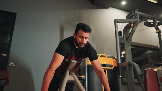 Low angle medium video shot of an athletic bearded man performing T-bar row machine exercises for upper back conditioning and lats muscle development in a gym.