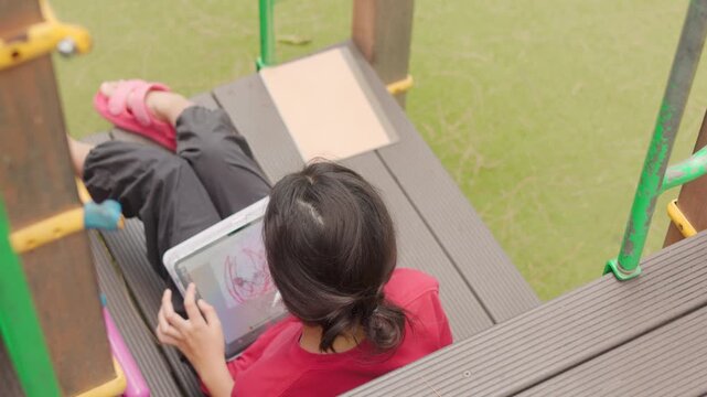 Top view of an Asian kid enjoying a relaxing hobby, drawing digital art on a tablet. Selective focus captures the elementary student spending casual leisure time at an outdoor school.
