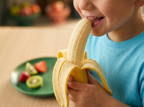 Child eating a banana at a table with fresh fruit nearby. Healthy eating, child nutrition and wellness. Fresh fruit snack and balanced diet. Healthy lifestyle and food choices.