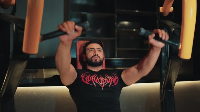 Low angle medium shot of a muscular bearded man performing incline chest press machine exercise for upper chest muscle hypertrophy and strength training in a gym.