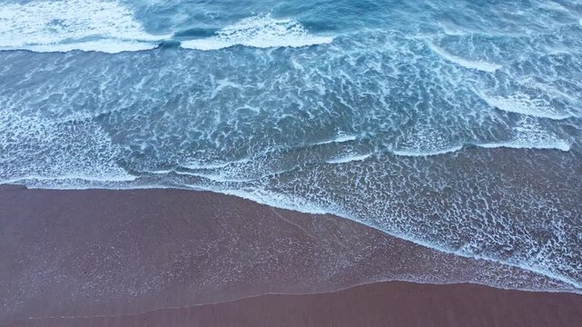 Aerial view of blue ocean waves washing over dark sand, creating layered white foam, wet shoreline textures, and elegant natural coastal patterns.