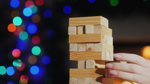 People playing wooden block game near fireplace with christmas lights background in cinematic warm evening setting.