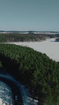 Drone footage of a winding road through dense pine forest beside a vast frozen lake on a clear winter day in the Ural Mountains, Russia, scenic wilderness landscape