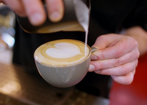 Cappuccino serving. Decorating drink. Barista hand making frothing hot cream heart pattern from pitcher to beverage coffee mug technique.