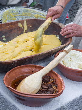 a man preparing traditional Romanian mămăligă (yellow cornmeal polenta) layered with white cheese and toppings in a large wooden trough at an outdoor cultural festival