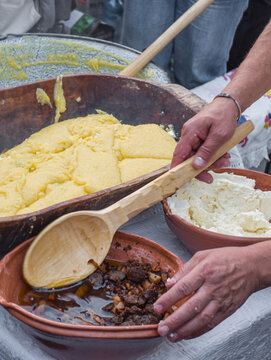 a man preparing traditional Romanian mămăligă (yellow cornmeal polenta) layered with white cheese and toppings in a large wooden trough at an outdoor cultural festival