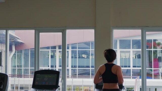 Rear view of an active Asian woman in her forties wearing a black sports bra while running on a treadmill. She exercises in a modern gym, looking out large windows during her daily cardio workout.