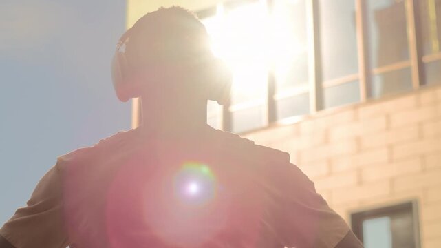 Happy African American man putting on headphones enjoying listening music in sunny city. Ethnic positive guy student optimistic male listen song audio in wireless earphones in sunlight sun holiday