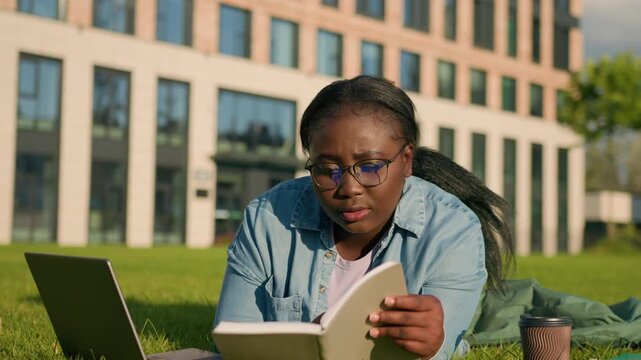 Concentrated African American woman studying learning reading notebook using laptop drinking coffee in park focused ethnic girl female read book preparing for exams course with computer lying on grass