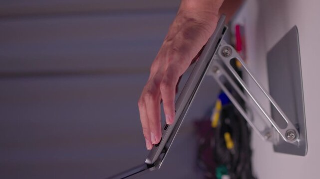 Close-up right hand adjusting tilted laptop mounted on adjustable metal hinge bracket with visible cables, reflecting soft light on aluminum housing in indoor workspace
