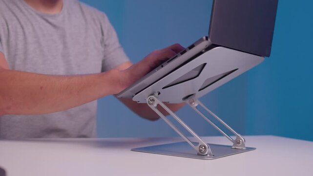 Close-up right hand adjusting tilted laptop mounted on adjustable metal hinge bracket with visible cables, reflecting soft light on aluminum housing in indoor workspace