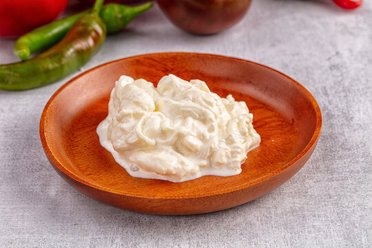 Wooden plate holding creamy stracciatella cheese with green chili peppers and red tomato in background on light gray surface