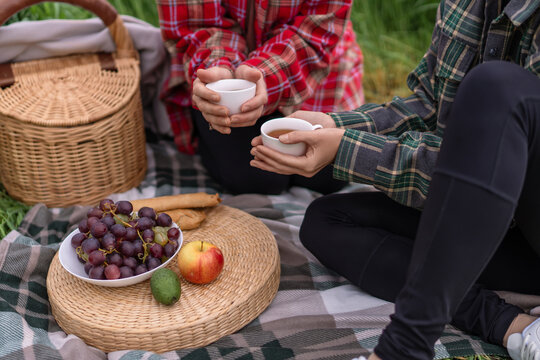 Autumn picnic friends holding hot tea mugs while sitting on blanket with fresh fruit and wicker basket