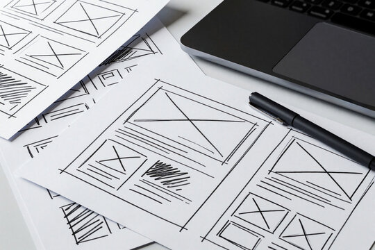 UX designer creates website wireframe sketches on paper. Hand drawn UI layout prototypes lie near modern laptop, black pen. Creative workspace for web development, app design, user experience