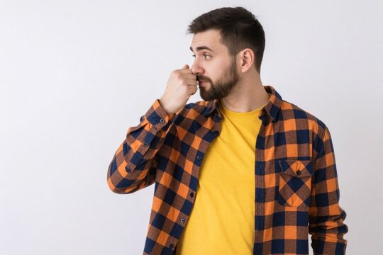 Young caucasian man with beard wears plaid shirt, yellow t-shirt. Holds nose with hand due to bad smell. Guy shows disgusted face reaction to unpleasant odor stinky aroma against white background.
