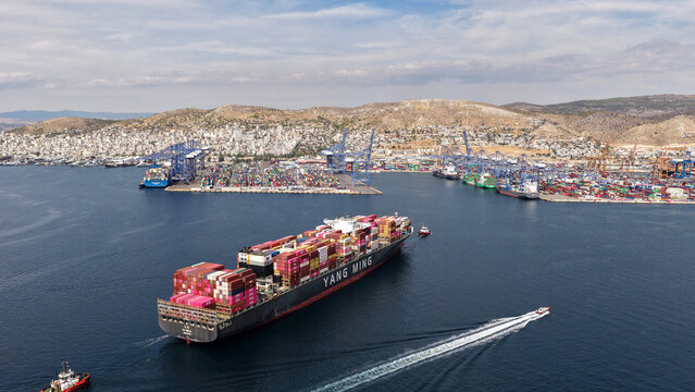 Aerial drone photo of truck size container cargo vessel approaching loading - unloading logistics port of Perama assisted by tug boat, Attica, Greece