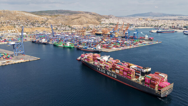 Aerial drone photo of truck size container cargo vessel approaching loading - unloading logistics port of Perama assisted by tug boat, Attica, Greece