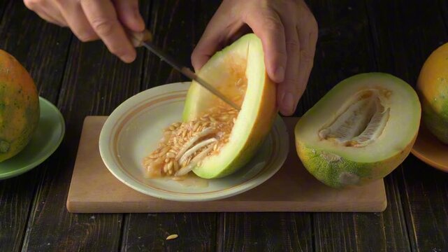 Person cutting ripe green melon and scraping seeds into a ceramic plate on a wooden cutting board, overhead food preparation scene in a rustic kitchen