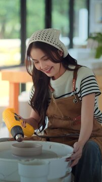 Young Asian woman using heat gun while making ceramic bowl in pottery studio, smiling female artisan working on handmade craft at pottery wheel, creative workshop.