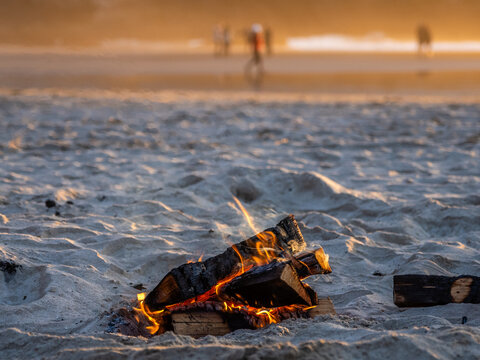 Small beach bonfire burning in the sand at Cannon Beach, Oregon, with warm sunset light, shallow depth of field, and blurred people walking near the shoreline in the background