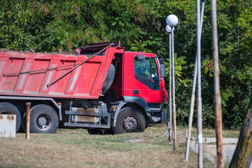 A red heavy-duty dump truck parked near a grassy area and green trees alongside park lampposts. © Andrey