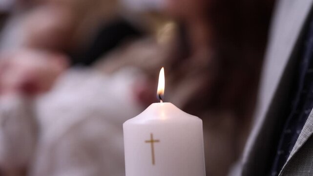 Close-up of a lit baptismal candle with a golden cross, softly blurred infant and priest's vestments in the background, capturing a quiet moment during a christening ceremony.