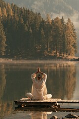 Fototapeta premium Rear view of a woman meditating crosslegged on a wooden pier over a calm forest lake at dawn for wellness background and meditation poster