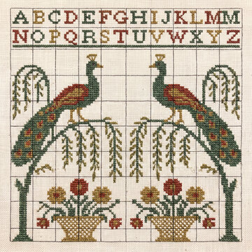 Vintage style cross stitch embroidery sampler featuring an alphabet grid with symmetrical peacock motifs and floral baskets in a traditional folk art design on textured linen fabric
