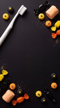 Vertical flat lay background with a white toothbrush, black and yellow mini pumpkins, marshmallows, and colorful wrapped candies arranged on a dark black surface. Halloween dental care.