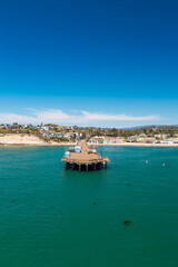 Fototapeta premium Aerial view captures Capitola Wharf extending into the ocean towards the sandy beach and coastal town buildings in Capitola, California, under a clear sky.