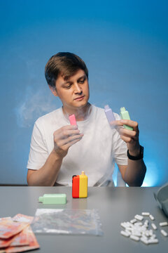 Drug dealer holding two colorful electronic cigarettes surrounded by smoke with more vapes, pills, money and plastic baggies on table against isolated blue background
