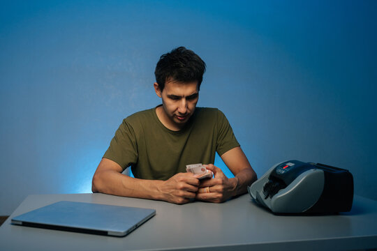 Portrait of cashier male counting banknotes with hand sitting at desk with laptop and automatic banknote counter machine at modern dark workplace, illuminated by soft blue light in background.