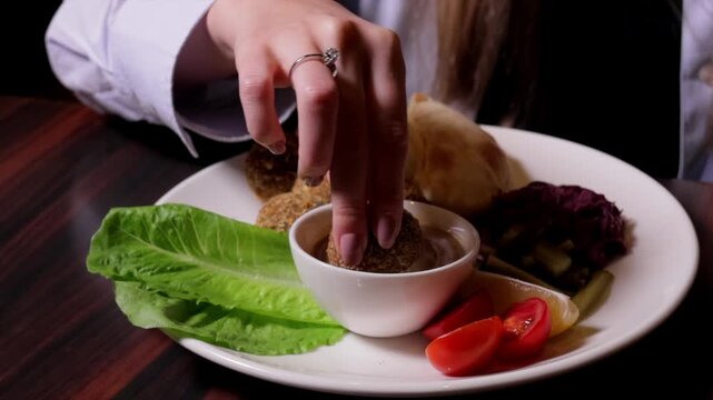 Close-up of hand dipping falafel beside hummus and pita bread