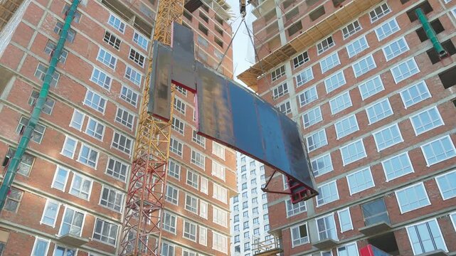 Crane lifting pallet of red clay bricks beside unfinished brick building at active construction site with workers, scaffolding, daylight sky and urban development context