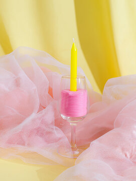 Tiny pink cake and yellow candle rest inside a wine glass amid pastel fabric. Great for stylish posters, promotional graphics and modern visual storytelling.