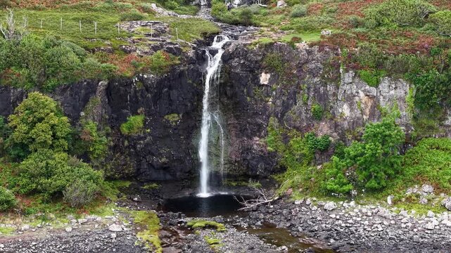 Coastal waterfall dropping off steep rock cliff face onto rocky shore on Scottish island, green bracken and ferns covering upper slopes around drop