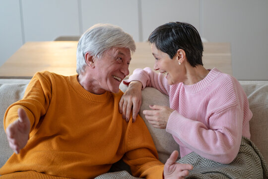 Smiling elderly husband and wife sit close together on couch, animated man gestures during amusing story, happy senior couple shares humor, communication and enjoyable retirement time at home.