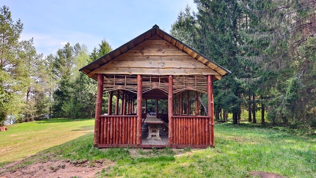 There is a wooden gazebo with a long wooden table and benches on the grassy lawn. There is a forest, a lake and a brazier nearby. Sunny spring weather and blue sky with feathery clouds
