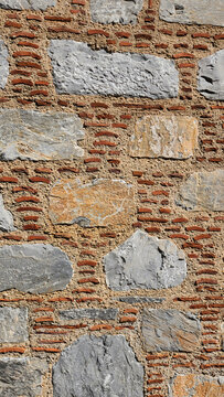 Traditional masonry wall built with irregular gray and yellowish natural stones, small red brick fragments arranged in horizontal bands, and sand-colored mortar, reflecting a construction style common